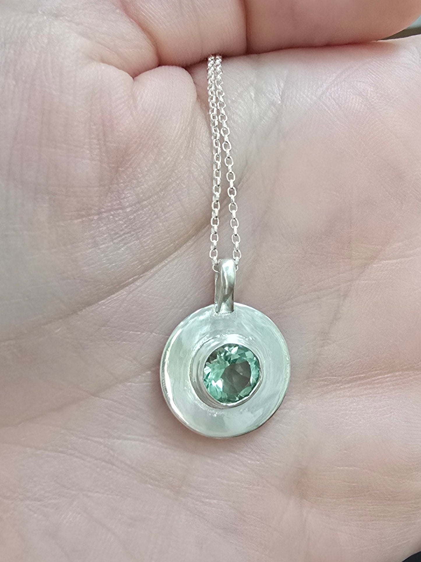 Hand holding a silver necklace with a green gemstone pendant.